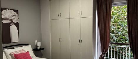 Un dormitorio con cama, armario, mesita de noche, un cuadro y una ventana con cortinas.