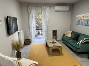 A modern living room with a green sofa, a wooden coffee table, a white armchair, and a view through a window with white curtains.