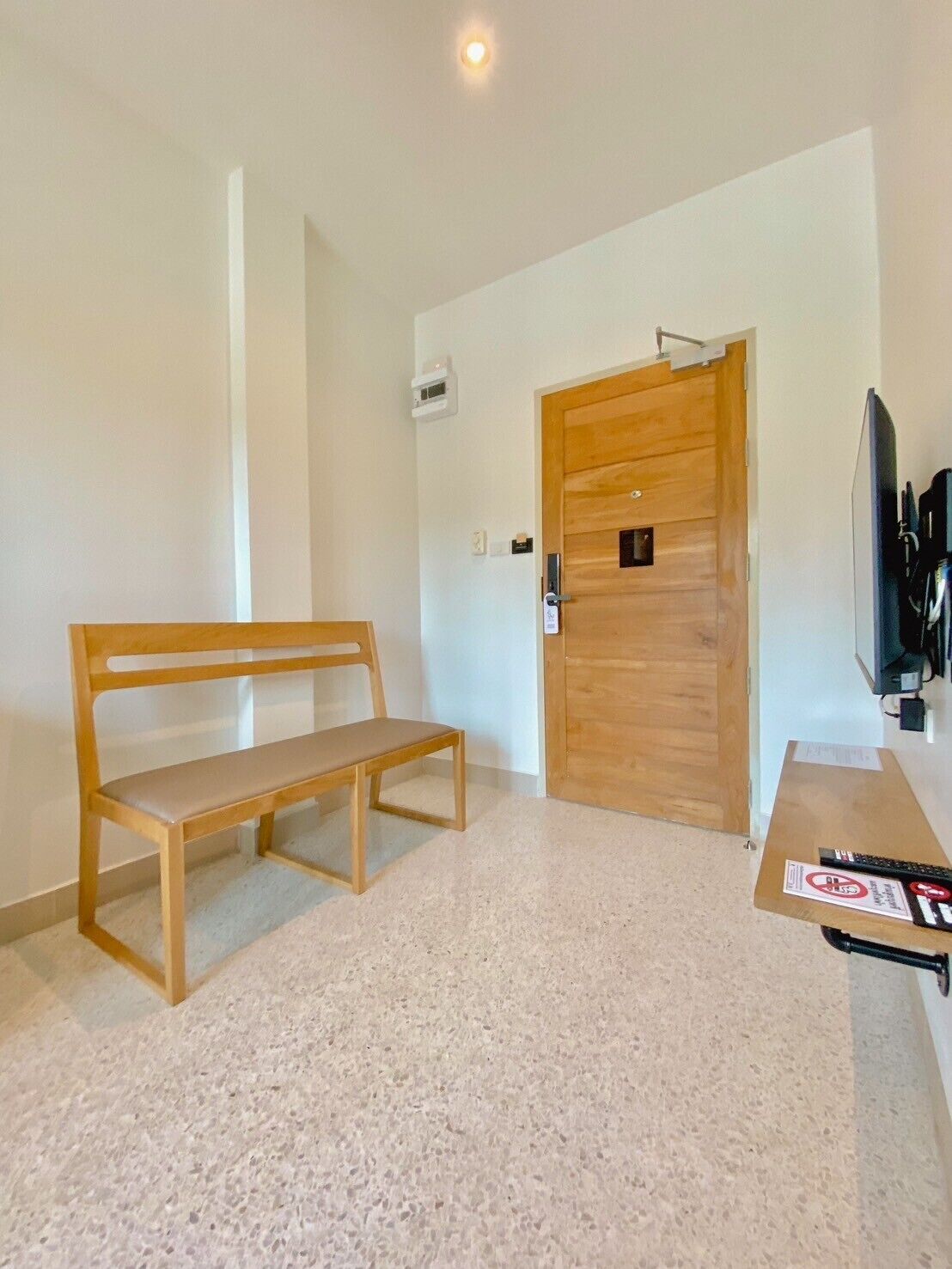 Una habitación con un banco de madera, una puerta de madera, un televisor y una mesita con un cartel.