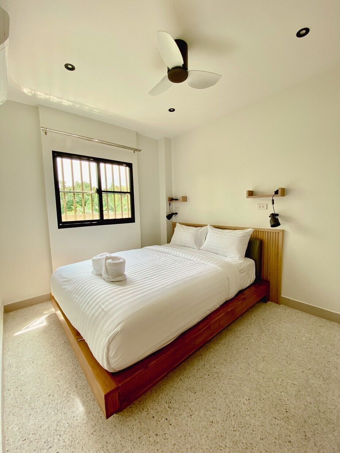 A bedroom with a bed, a ceiling fan, a window, and a nightstand with a lamp.
