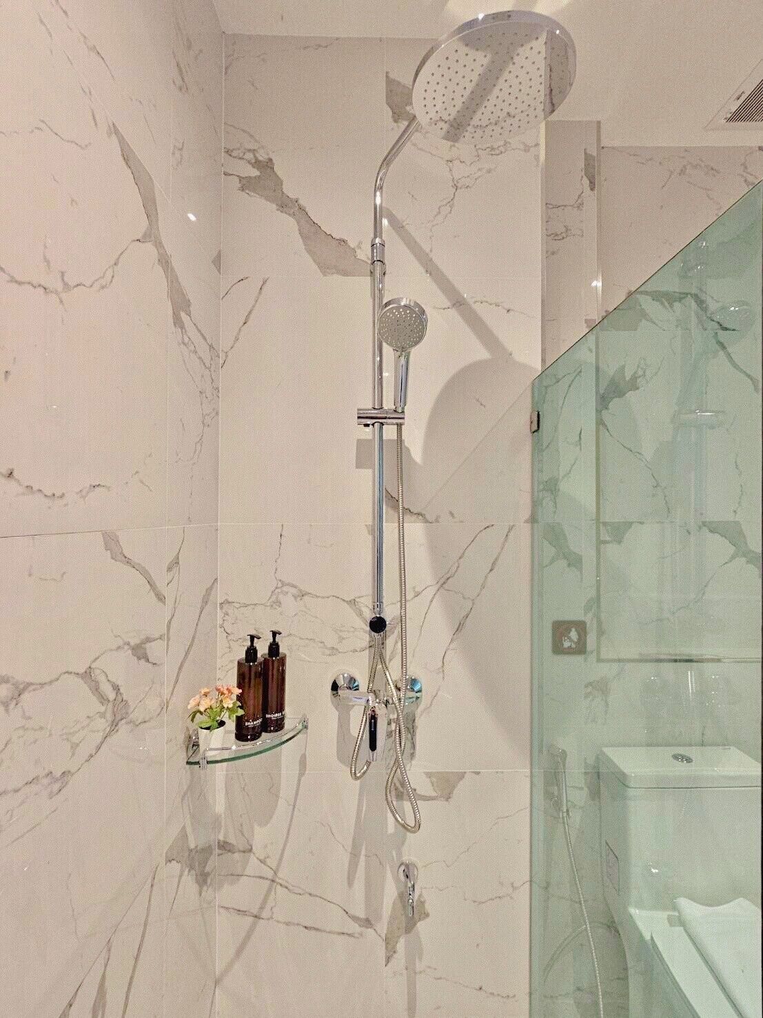 Un baño con ducha, paredes de mármol, un separador de vidrio y artículos de tocador en un estante.