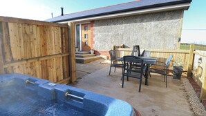 A hot tub in a backyard with a wooden fence, a patio table with chairs, and a house with a stone exterior.