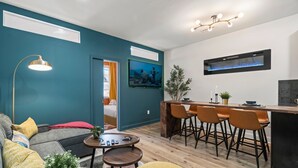 A modern living room with a sofa, coffee table, and a TV. There is a kitchen area with a bar and stools.