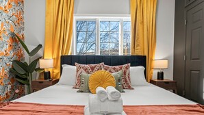 A neatly made bed with a white duvet, a brown throw blanket, and rolled towels in front. There are two bedside tables with lamps, a large window with yellow curtains, and a zebra-patterned wall.