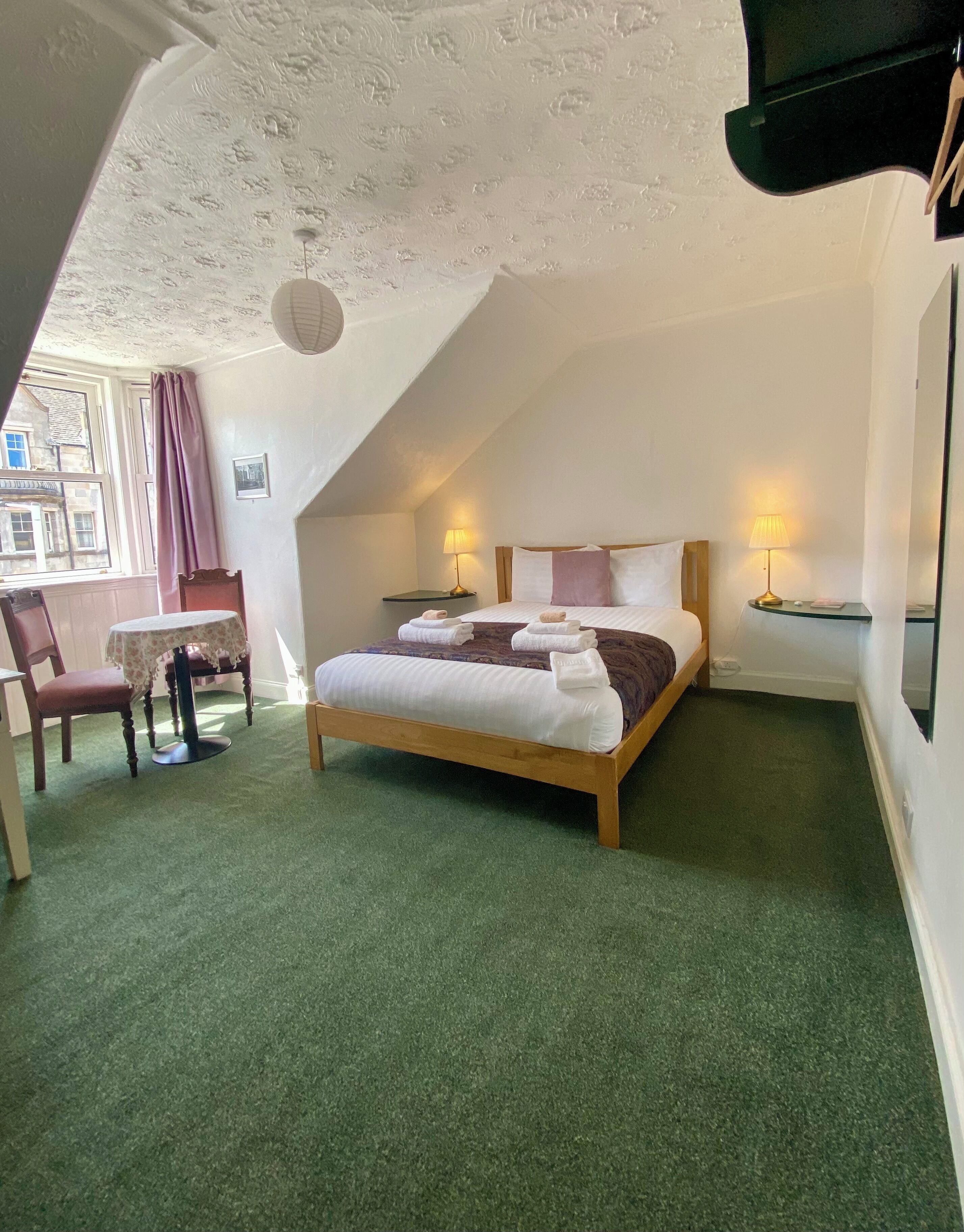 A bedroom with a bed, two bedside tables, a chair, a small table, and a window with curtains.