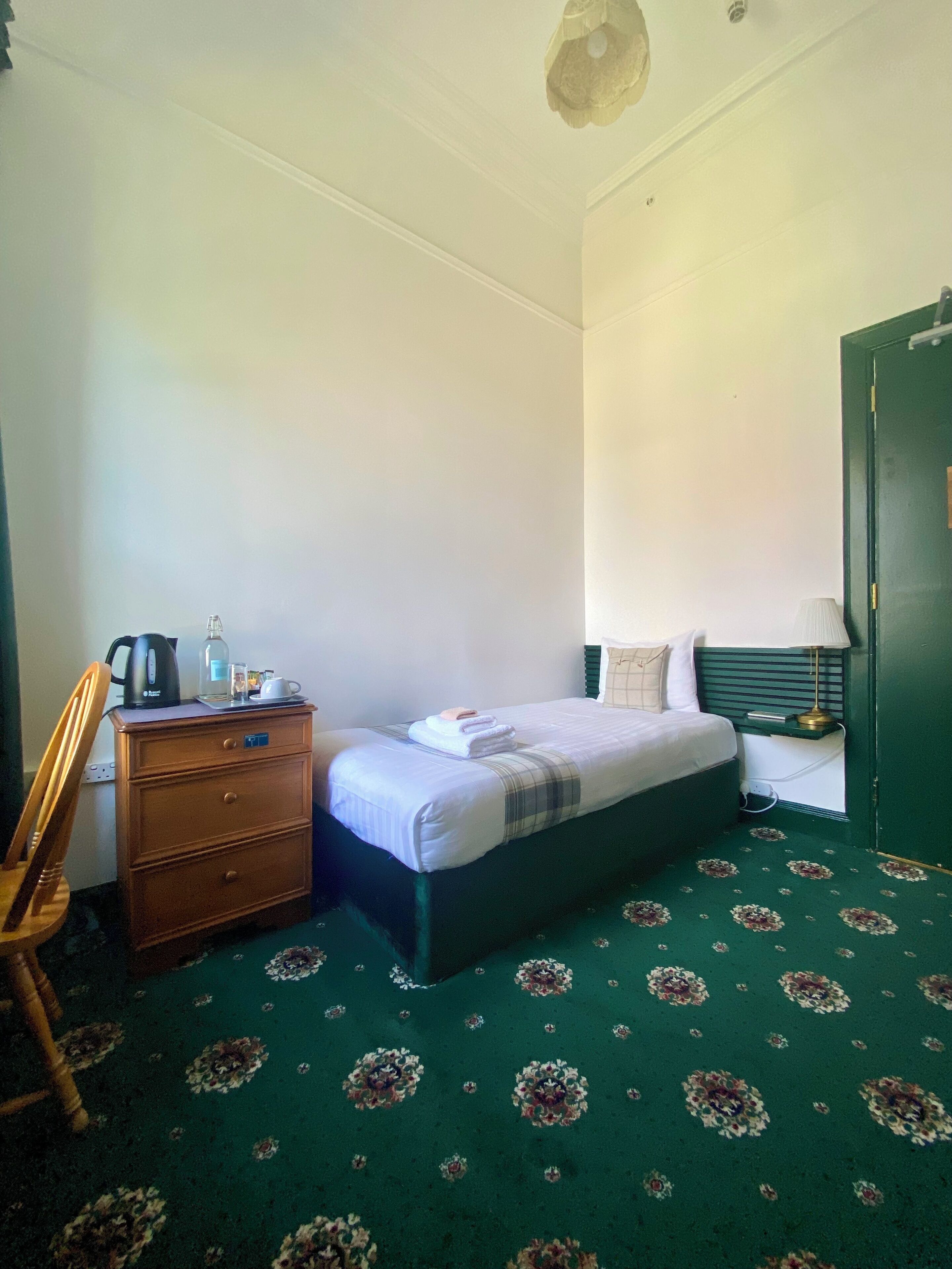 Un dormitorio con una cama, un aparador, una silla, una lámpara y una alfombra verde con motivos florales.