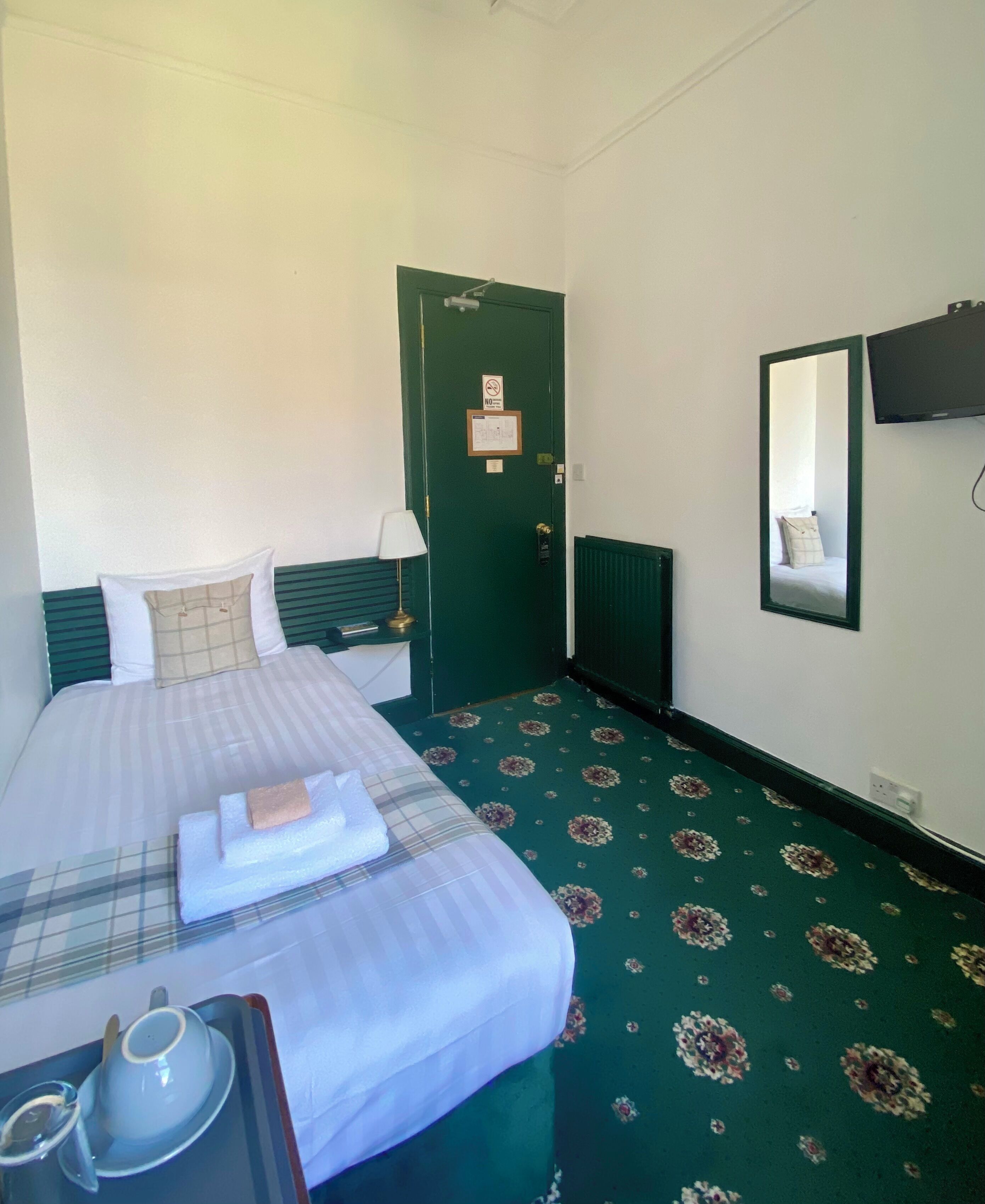 A hotel room with a bed, a nightstand, a mirror, a green carpet, and a door with a green frame.