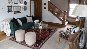 A living room with a green sofa, ottomans, a coffee table, and a patterned rug. A staircase is visible in the background. - Lena's Place (Findlay)