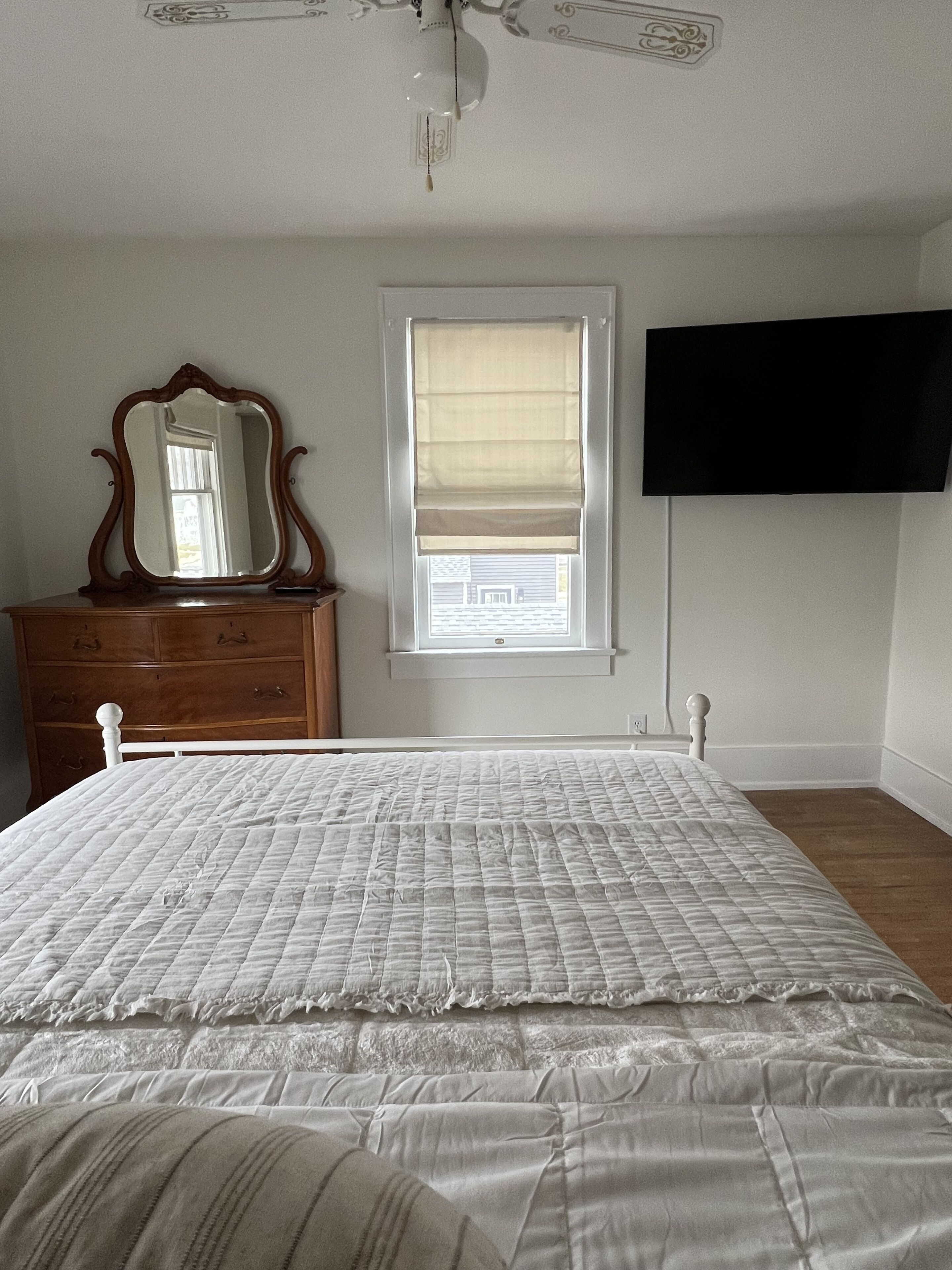 Een slaapkamer met een bed, dressoir, spiegel, raam met zonwering en een aan de muur bevestigde televisie.
