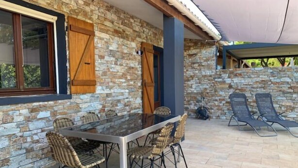 A stone building with a glass table and wicker chairs, a covered patio area, and two lounge chairs.