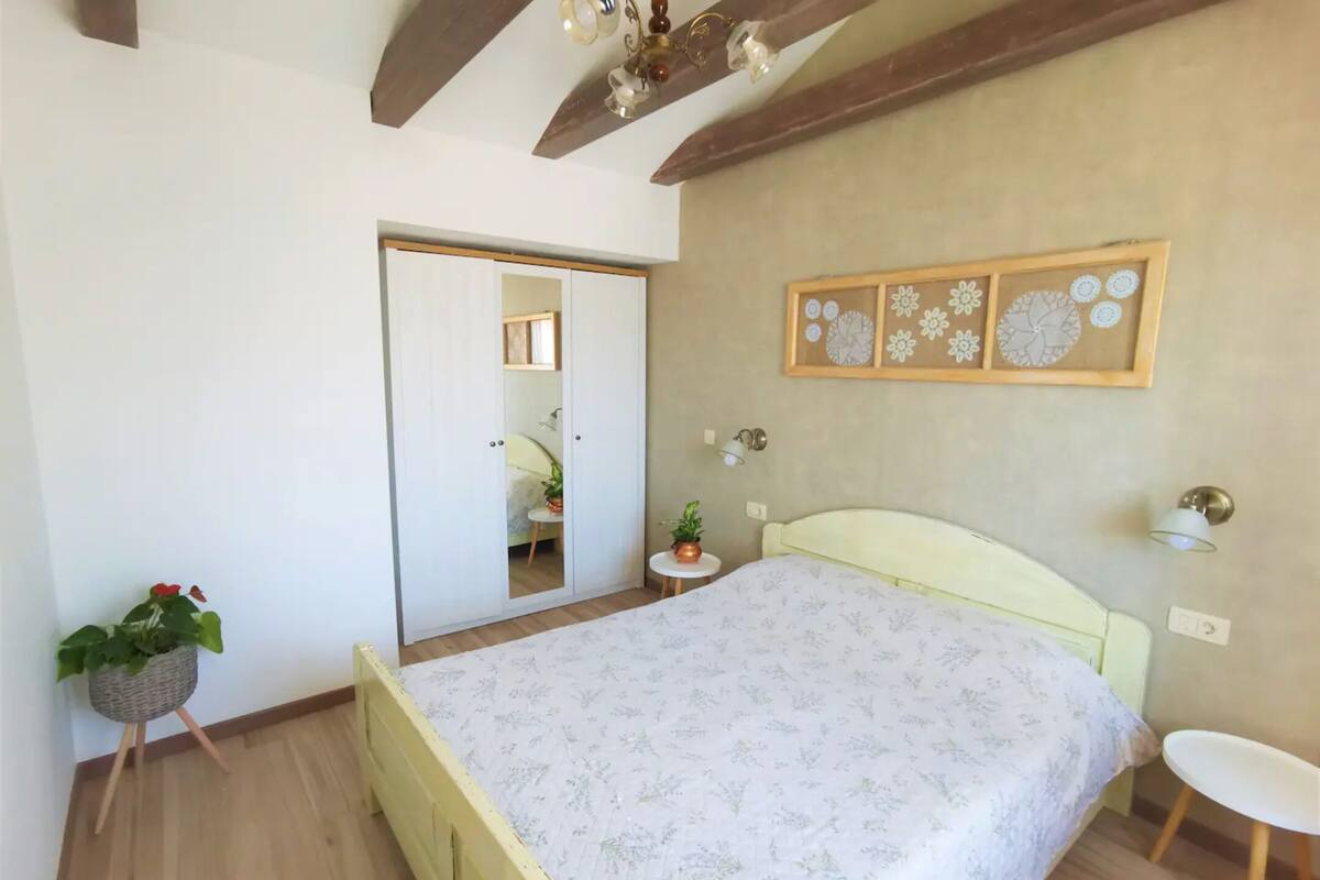 A bedroom with a bed, a small bedside table, a potted plant, a wall-mounted light fixture, and a ceiling fan.