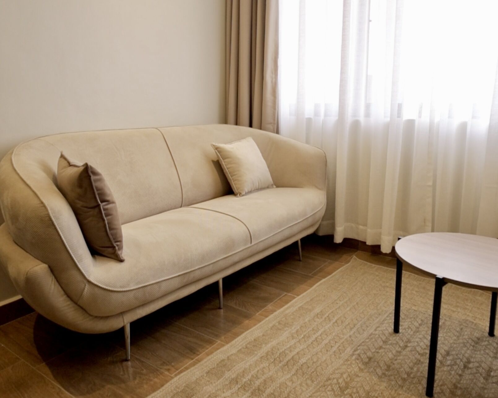 Un sofá beige con dos almohadones, una mesita redonda y un tapete estampado en una habitación con ventana y cortinas.
