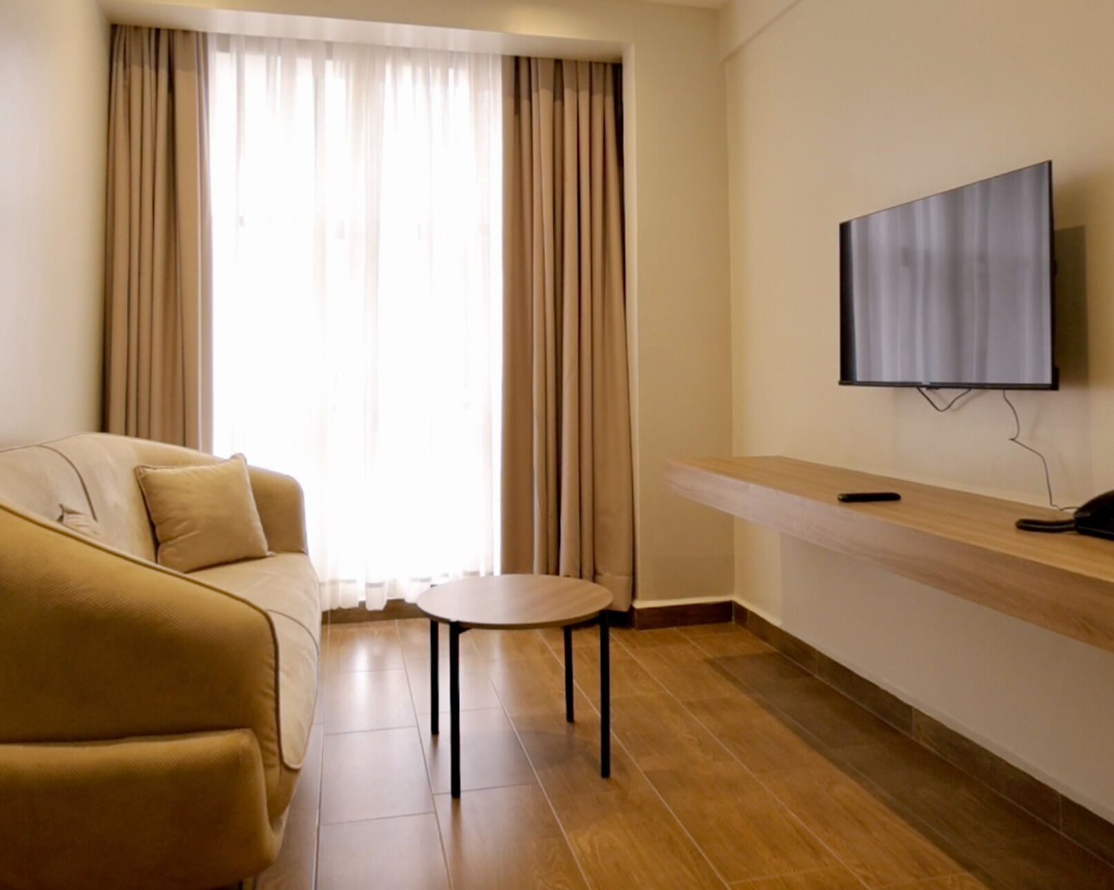 A modern living room with a beige sofa, a small round table, and a flat-screen TV mounted on the wall.
