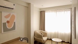 Una habitación de hotel moderna con una mesa de comedor de madera, un sillón color beige, una pequeña mesa redonda y un gran ventanal con cortinas translúcidas.