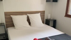 Een net opgemaakt bed met een donkere spreidek en twee kussens, een klein nachtkastje met een lamp, en een geruit vloerkleed.