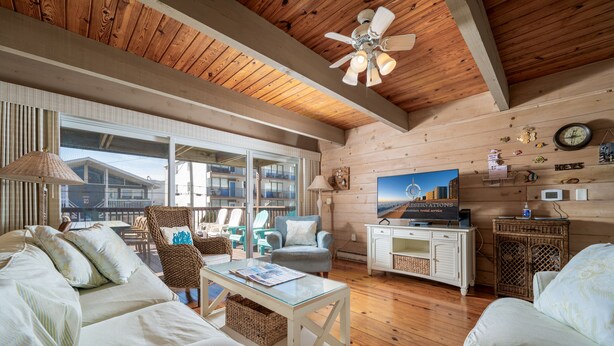 A cozy living room with a wooden ceiling, a large window, wicker furniture, and a flat-screen TV. - Oceanblock Townhome Mid Town with Pool (Ocean City)