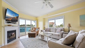 Ein Wohnzimmer mit Kamin, Fernseher, Deckenventilator, Sofa, Couchtisch und großen Fenstern.
