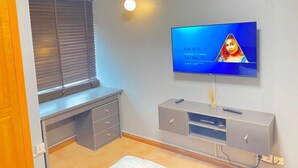 A modern living room with a flat-screen TV mounted on the wall, a grey entertainment unit below, a window with blinds, and a wall-mounted air conditioner. - Mulliner Towers (Lagos)