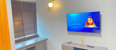 A modern living room with a flat-screen TV mounted on the wall, a grey entertainment unit below, a window with blinds, and a wall-mounted air conditioner.