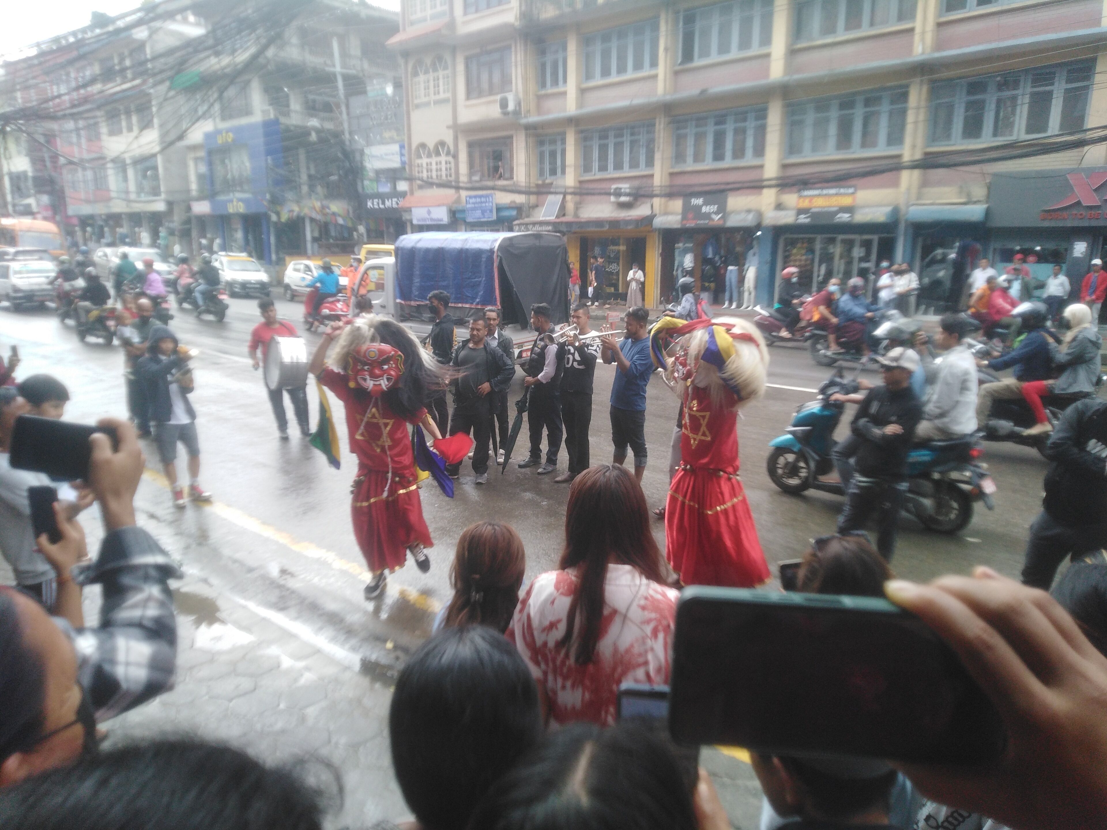 A street scene with a crowd of people, some holding cameras, and a few individuals in red costumes.