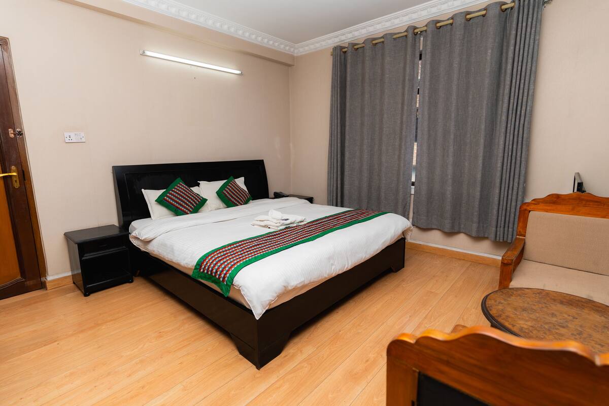 A neatly made bed with a white and green striped blanket, a wooden nightstand, a small table, and a chair in a room with wooden flooring and grey curtains.