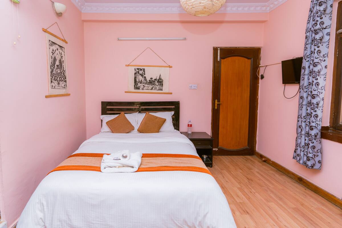 A neatly made bed with a white and orange bedspread, a wooden headboard, and two brown pillows. There is a small nightstand with a lamp and a bottle of water. The room has pink walls, a wooden floor, and a door with a curtain.