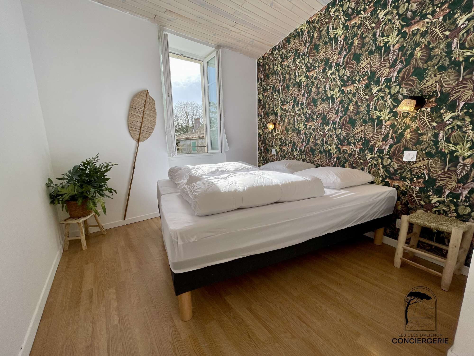 Una camera da letto con un letto, una finestra, un muro decorato con un motivo a foglie, una pianta su uno sgabello e un tavolino con una lampada.