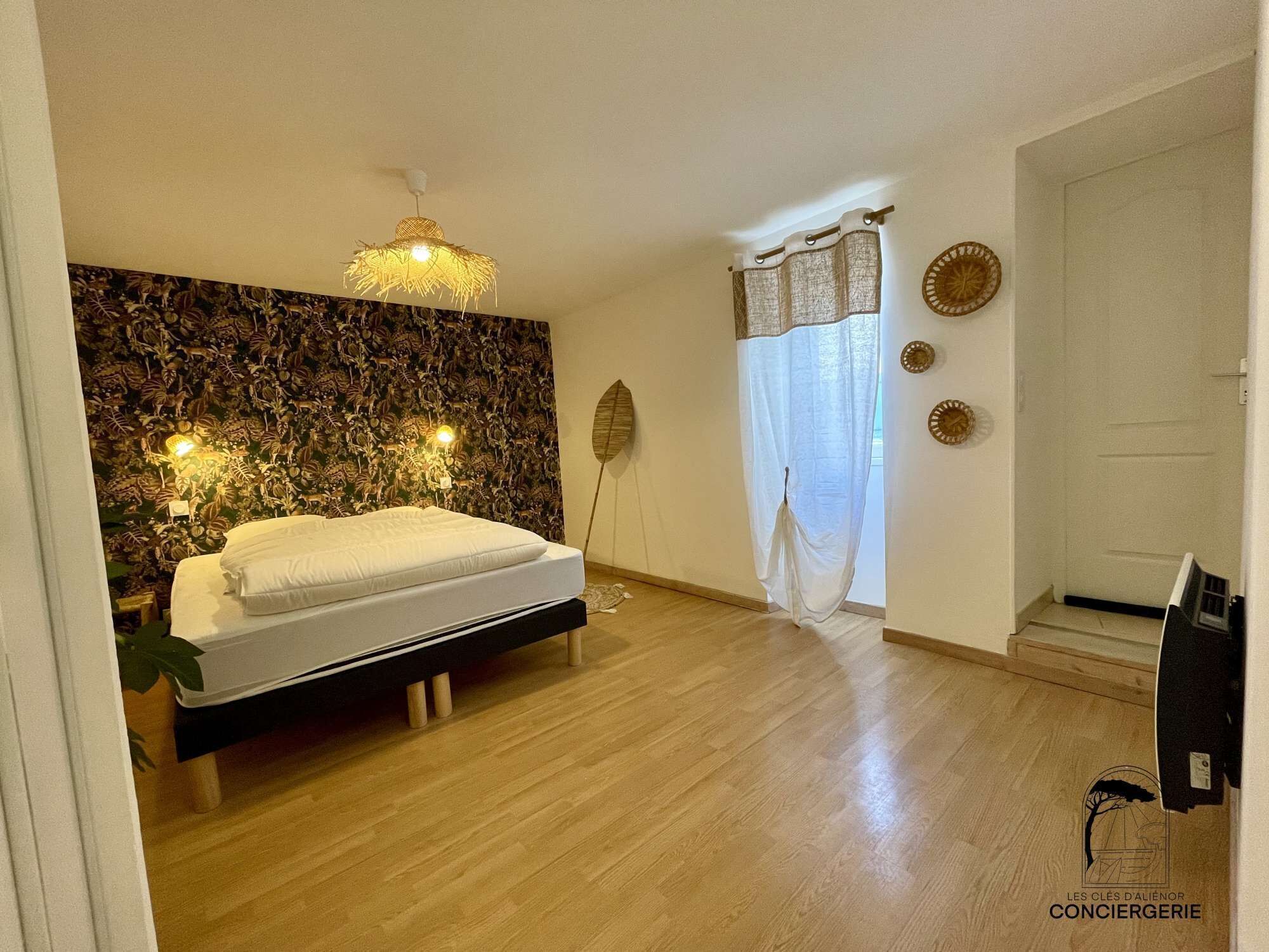 Une chambre à coucher comprenant un lit, un luminaire fixé au mur, un mur décoratif, une fenêtre avec des rideaux et une porte.