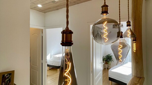 A modern interior with exposed wooden beams, a hanging light fixture with a spiral filament, and a glimpse of a bedroom with a bed and a plant.
