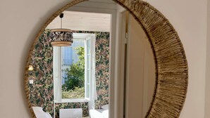 A round mirror with a woven frame reflecting a room with a bed, a small table, and a window with a view of greenery outside.