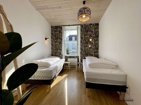 Una habitación pequeña y moderna con dos camas individuales, techo de madera, un ventanal con vistas a edificios y una lámpara colgante.