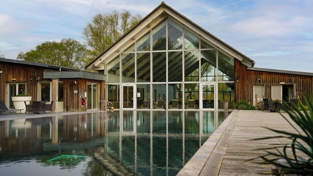 A modern building with large glass windows and a pool area. - Eider Cottage: Lakeside Escape with Spa Access (Cirencester)