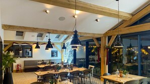 A restaurant with wooden beams, hanging pendant lights, and a large window with a city view. Tables are set with glasses and cutlery. - Eider Cottage: Lakeside Escape with Spa Access (Cirencester)