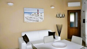 Un coin repas avec une table et des chaises blanches, un tableau représentant un phare accroché au mur et une porte en bois.