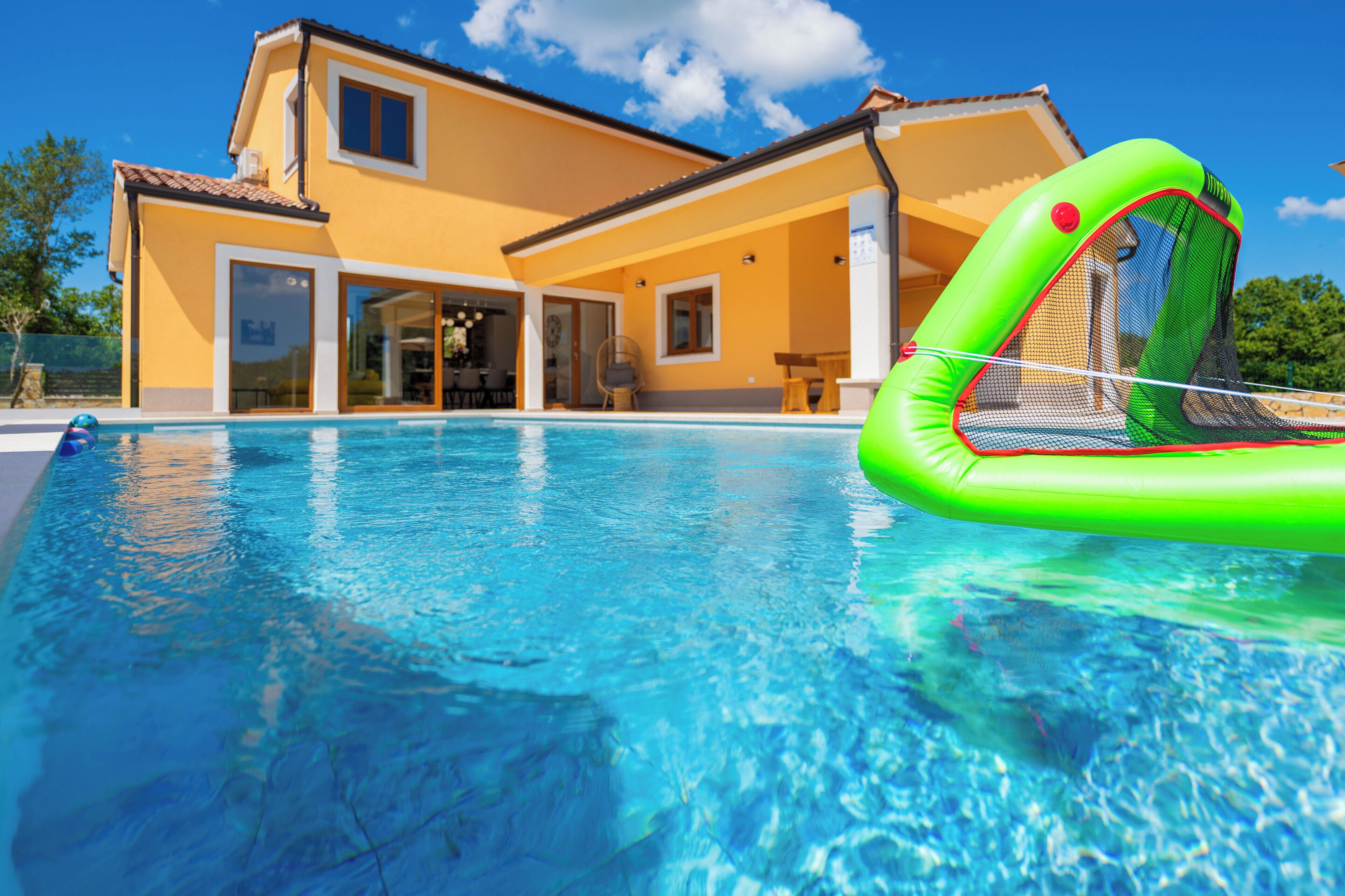 A swimming pool with a green inflatable float, a yellow house with large windows, and a clear blue sky.