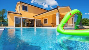 A swimming pool with a green inflatable float, a yellow house with large windows, and a clear blue sky.
