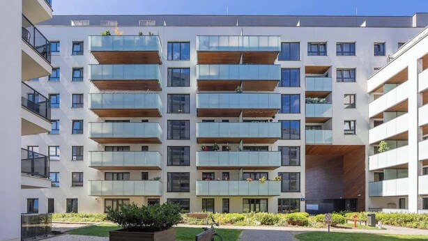 Un moderno edificio residencial de varios pisos con balcones, un patio con un banco y una pequeña área ajardinada.