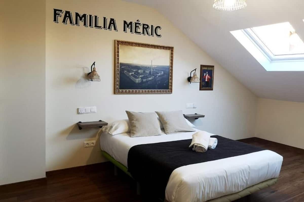 Quarto com cama, claraboia, quadro emoldurado e luminária na parede.