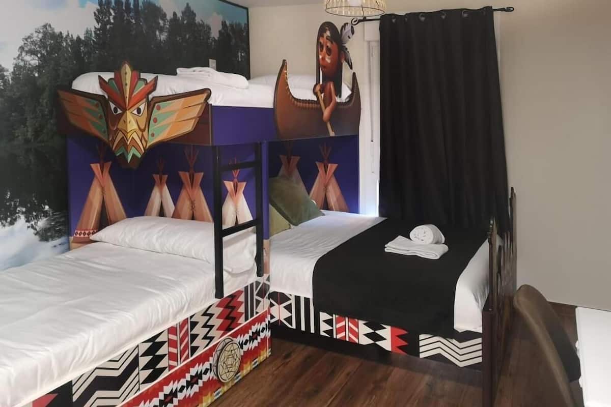 Quarto com duas camas, uma com cabeceira colorida e outra com padrão geométrico em preto e branco. Há um quadro na parede, um lustre e uma cortina preta.