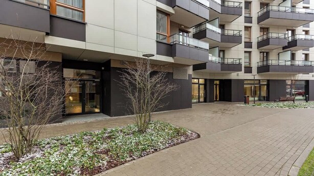 Un edificio moderno de varios pisos con techo plano, ventanas amplias y un patio empedrado con un pequeño jardín.
