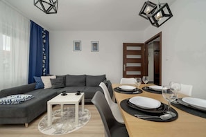 A modern living room with a grey sofa, a small white table, and a dining area with a wooden table set for two.