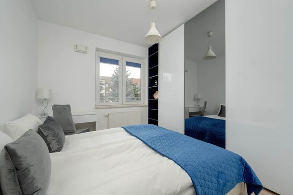 A modern bedroom with a comfortable double bed, soft gray and blue bedding, and a large mirror reflecting natural light.