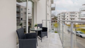 A balcony with a glass railing, two wicker chairs, a small round table with a plant and a mug, and a view of neighboring buildings and a parked car.