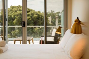 A hotel room with a large window, a bed, a lamp, and a view of trees and a building.