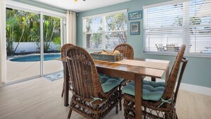 Comedor con sillas de mimbre y una mesa de madera, ventanas amplias con persianas y vista a una piscina.