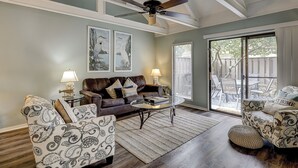 A living room with a brown leather sofa, patterned armchairs, a coffee table, and a ceiling fan. There are two wall-mounted lamps and a large window with blinds.