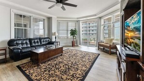 Un salon avec un canapé en cuir noir, une table basse en bois, un tapis à motifs, un ventilateur de plafond et un grand écran de télévision.