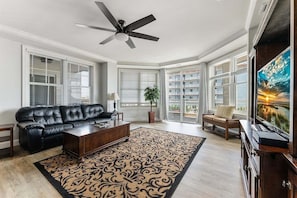 A living room with a black leather sofa, a wooden coffee table, a patterned rug, a ceiling fan, and a large TV screen.