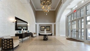 Un hall d’entrée spacieux avec un haut plafond, un lustre, un comptoir de réception, un espace salon et une grande porte d’entrée en verre.