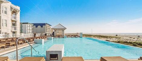 En swimmingpool med solstolar, en byggnad med balkonger och en strand i bakgrunden.